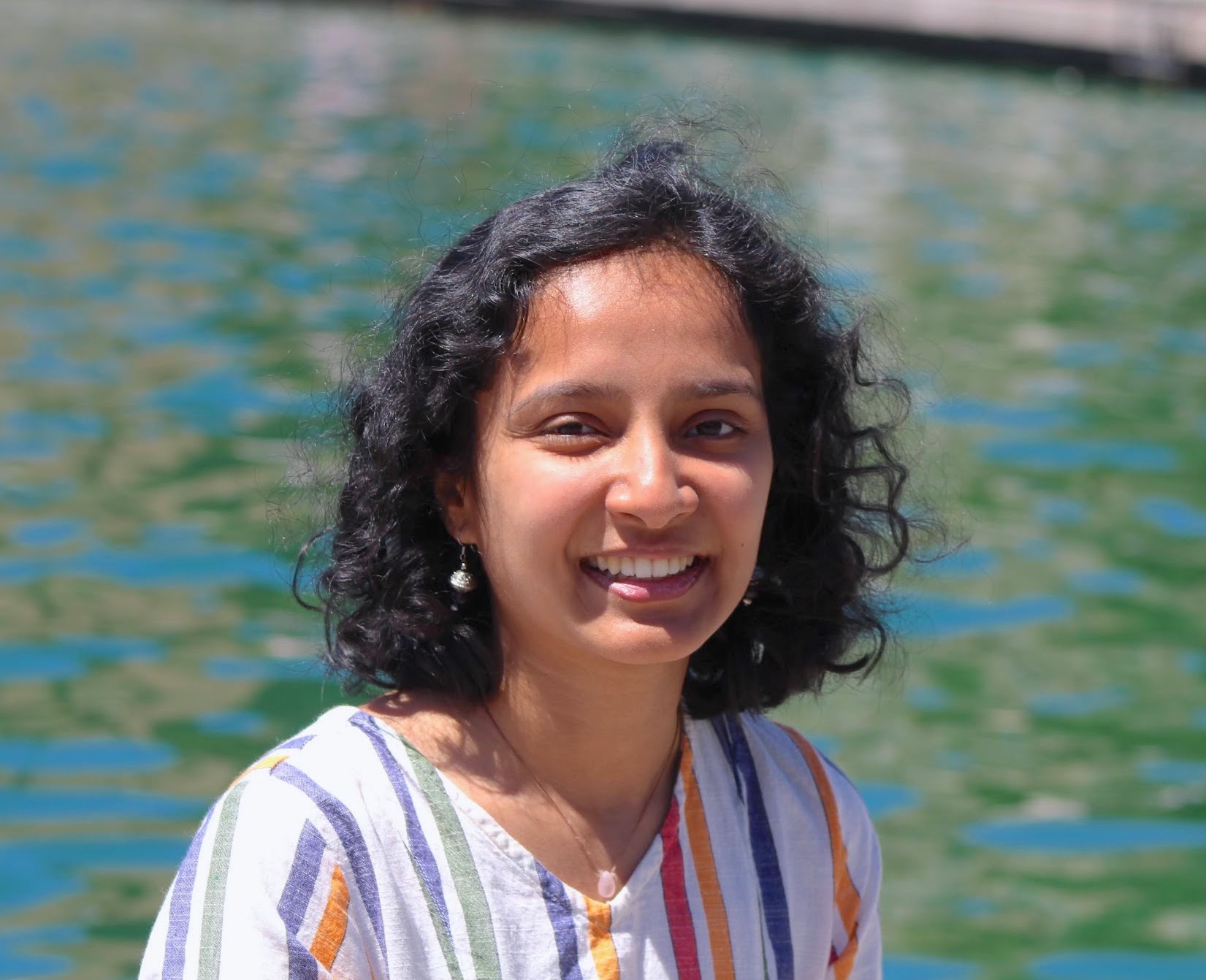 Aashaka, an Indian woman with short curly black hair, smiling with bluegreen water in the background.